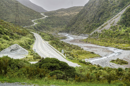 A scenic mountain road in Arthurs Pass New Zealand winds through a green valley beside a rocky riverbed, framed by lush hills and power lines, creating a dramatic natural landscapeの写真素材
