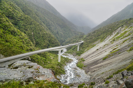 Otira Viaduct Lookout Arthurs Pass, bridge crosses a rugged gorge between green mountains, with a rushing river below and misty peaks in the distance.の写真素材