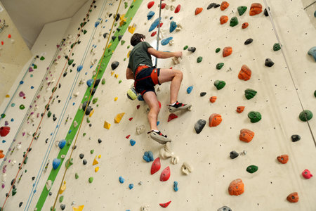 man with safety rope and alpine harness climbing indoor wall with colorful holdsの写真素材