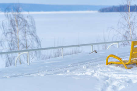 Winter photography. Sunny day. Bright yellow park bench. The river is covered with ice, covered with snow. A big tree.の写真素材