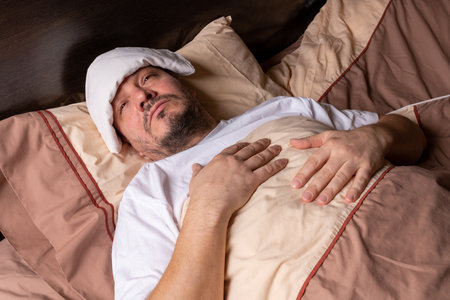 The young man feels unwell and lies in bed. Takes antipyretic tablets to reduce fever. Body temperature monitoring to track sickness.の写真素材