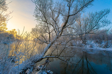 Experience the awesome-inspiring beauty of nature's winter magic. Watch as the sun sets and colors the sky in fiery shades of orange and pink. See the warm glow of the sun reflected in the icy river.の写真素材