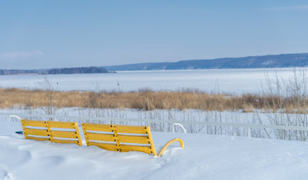 Capture the beauty of winter in a photo A sunny day illuminates a park bench A river covered in ice and snow creates a serene atmosphereの写真素材