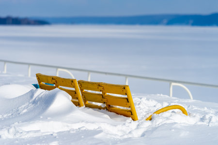 Capture the beauty of winter in a photo A sunny day illuminates a bright yellow park bench A river covered in ice and snow creates a serene atmosphereの写真素材