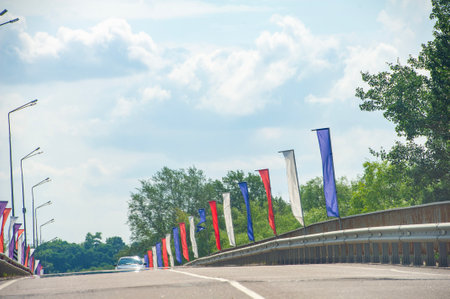 A modern bridge lined with red, white, and blue flags stretches across a highway. The vibrant colors contrast against the sky and trees, adding a festive touch to the journey.の写真素材