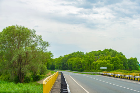 A well-maintained road with bright yellow guardrails, curving gently into dense green forests. The sky is slightly overcast, adding a calm and serene mood to the landscape.の写真素材