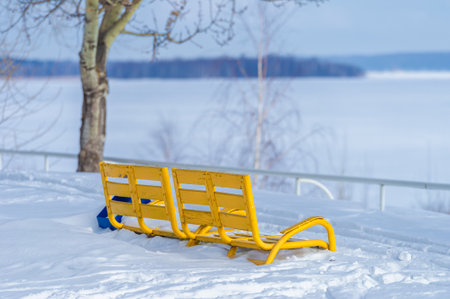 Capture the beauty of winter in a photo A sunny day illuminates a bright yellow park bench A river covered in ice and snow creates a serene atmosphereの写真素材