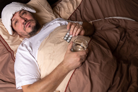 Man in bed with a glass of water and pills in his handの写真素材