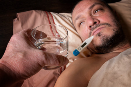 The young man feels unwell and lies in bed. He takes antipyretic pills to bring down his...