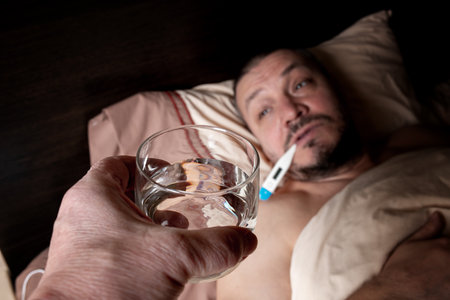 The young man does not feel well and is resting in bed. He takes antipyretic pills to bring down his temperature. He monitors his body temperature to track the progress of his illness.の写真素材