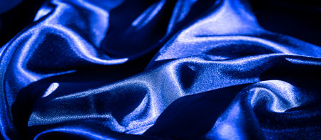 Blurred blue satin fabric flowing smoothly with soft undulating waves. The shiny material reflects light, enhancing its richness, luxurious deep blue hue and creating a dynamic surface.の写真素材