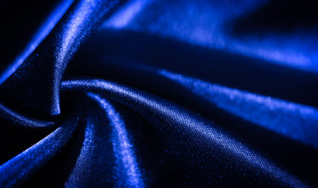 Blurred close-up of deep blue fabric with elegant folds and subtle twists. The smooth, glossy surface reflects light, creating a luxurious and fluid texture, perfect for abstract textile designs.の写真素材