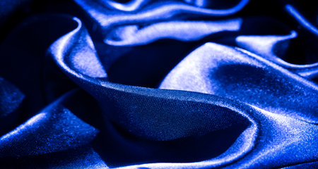 Blur of shiny blue satin fabric in smooth, fluid folds. The soft, glossy texture reflects light, highlighting the luxurious, silky material and creating elegant curves throughout the deep blue fabric.の写真素材