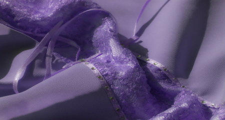 A blur of lavender lace fabric with sheer sections featuring delicate ribbon embellishments. The fabric folds gracefully with soft textures highlighted by shimmering floral lace.の写真素材