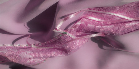 A blur of shimmering pink lace fabric with intricate floral and mesh patterns, with a delicate, sparkling stripe lined with small green gemstones. The texture adds elegance.の写真素材