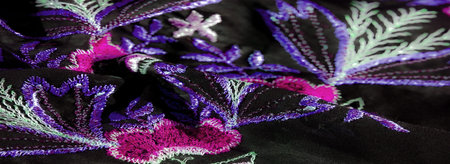 Blur of dark fabric with vibrant pink and purple embroidered flowers, accented by green leafy stems....