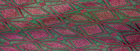 Blurred close-up of vibrant green fabric with intricate magenta geometric embroidery. The shimmering...