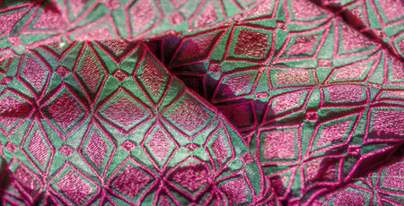 Blurred close-up of a green fabric with intricate geometric pink embroidery, displaying smooth folds...
