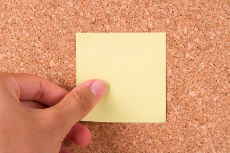 Hand and blank, yellow sticky post it note for your message on cork bulletin board.の写真素材