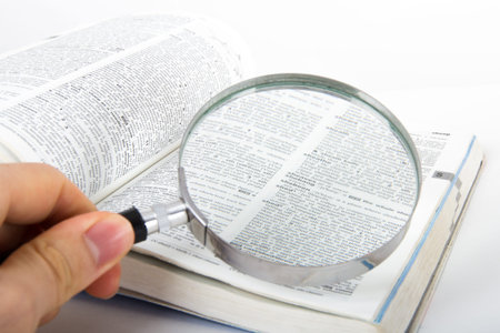 Hand holding classic style magnifying glass, searching for a word on open dictionary book, isolated...