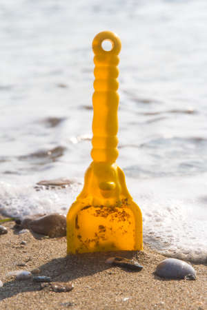 Yellow toy scoop standing on sand near sea with foam.の写真素材