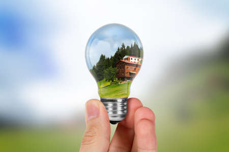 Energy saving concept, hand holding light bulb with green landscape and blue weather inside.の写真素材