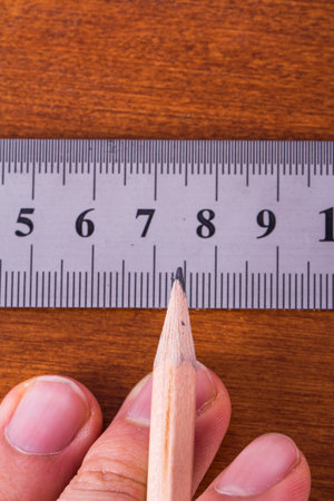 Macro view of hand holding wood pencil and steel ruler for engineering drawing on wooden table.