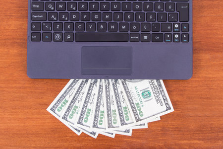 Detail view of laptop and one hundred dollar banknotes on wooden table.
