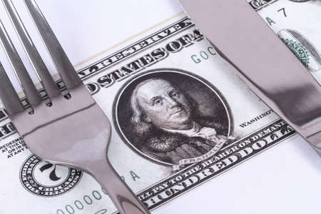Close up detailed view of one hundred dollar banknote under a steel fork and knife, isolated on white background.の写真素材