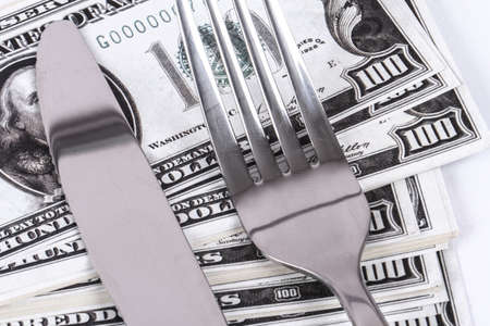 Close up detailed view of one hundred dollar banknotes under a steel fork and knife, isolated on white background.の写真素材