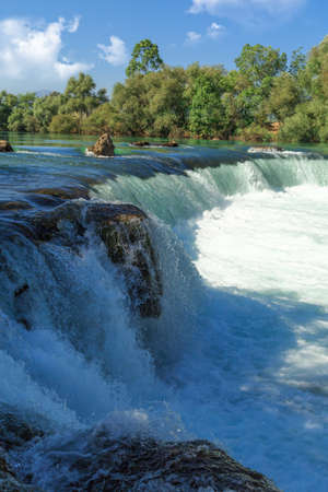 View of flowing Manavgat Waterfall in Antalya, Turkey, with green trees around, on cloudy blue sky background.の写真素材