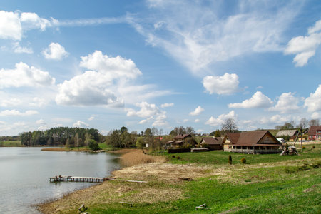 View of small wooden houses on lakeside among trees, on cloudy blue sky background.