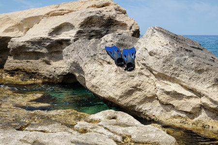 A pair of blue snorkel fins and a mask are casually placed on a sunlit rock by the shoreline, with clear turquoise water gently lapping at the rocks.の写真素材