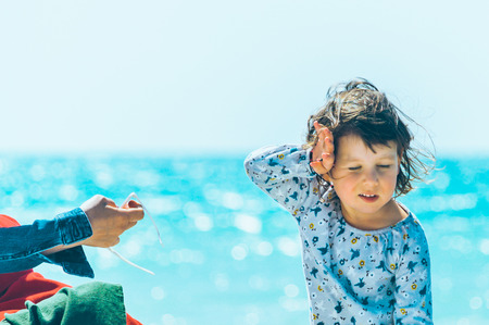 Three year old girl hiding her face from the wind. Mother giving her glasses. The sea at background.の写真素材