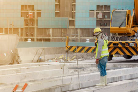 The construction worker in the green waistcoat and the yellow helmet and a black mask standing  on the steel beam in the construction area with yellow crane in the background.の写真素材