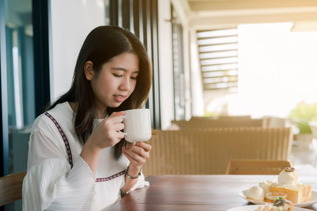 Asian beautiful woman Wearing a white shirt, sitting, drinking hot coffee In the bakery Happily...