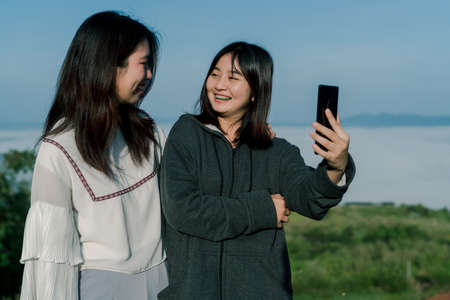 Two Asian girls wearing a sweater, take a selfie phone, take pictures in the tourist area behind the fog and mountains with a smiling expression of happiness..の写真素材