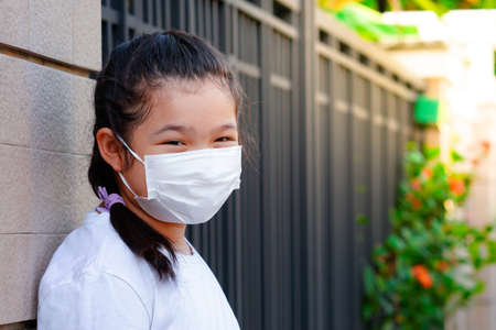 Portrait: Half-body of an Asian girl in a white shirt with a medical mask on her mouth, leaning against the wall of the house.の写真素材