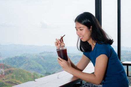 A beautiful long-haired Asian woman in a blue shirt sits in a coffee shop with a beautiful mountain...