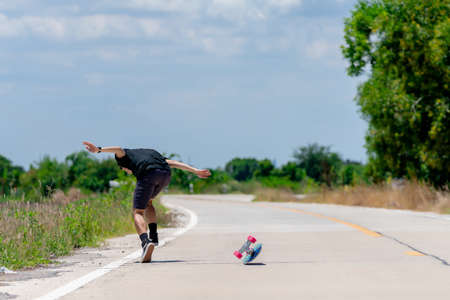 A young Asian man is wearing a black shirt and pants. play skateboard show an accident On a country road on a sunny day with sky. Looking at the camera, Play surf skate.の写真素材