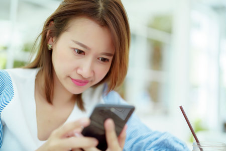 Portrait A beautiful, white Asian woman in a white shirt is happily sitting on her phone in a bakery.の写真素材