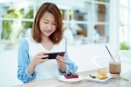 Portrait A beautiful, white Asian woman in a white shirt is happily sitting on her phone in a bakery.の写真素材