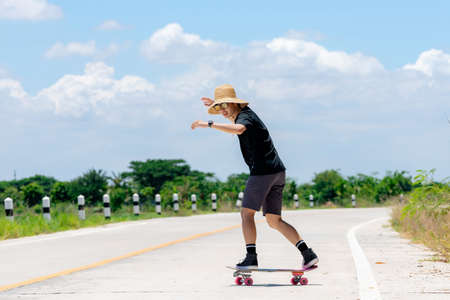 A young Asian man wearing a black shirt and pants, wearing a straw hat, is playing skateboarding. and show style On a country road on a sunny and sunny day., Play surf skateの写真素材