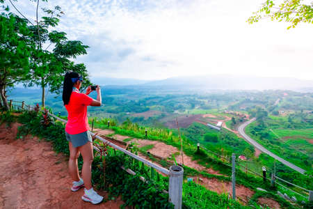 Female trail runners, wearing pink runners, sportswear, standing for a view with a mobile phone on a high mountain with a happy mood, on a clear dayの写真素材