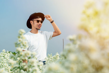 A young Asian man in a white shirt in a black hat stands posing in a white flower garden on a clear...