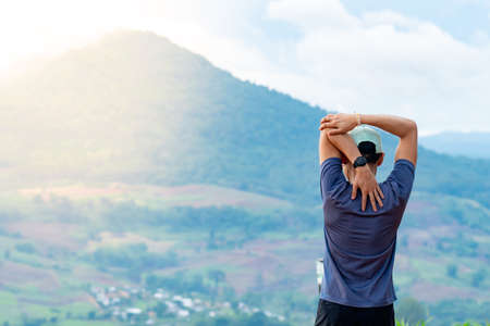 Half-body shot of an Asian male runner in a blue shirt and hat standing warm up in preparation for a jogging run. It is located on a high cliff with beautiful mountains in the background.の写真素材