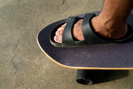 men's feet In sandals, stepping on a black Surf Skate on the sand in the park.