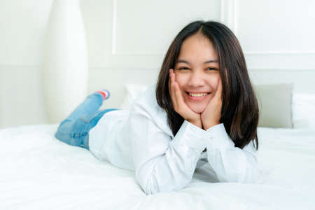 Portrait Cute Asian teen with long hair Wearing a white shirt and lazing around in bed happily in the bedroom.の写真素材