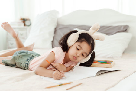 4k 50fps Cute little girl having fun in cute kindergarten age. Use colored pencils to draw a picture on the paper. Happily put the rabbit headphones on the bed. Concept family and happy learningの写真素材