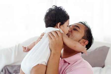 Asian single father playing with his 2 month old son happily holds him up and kisses his cheek....